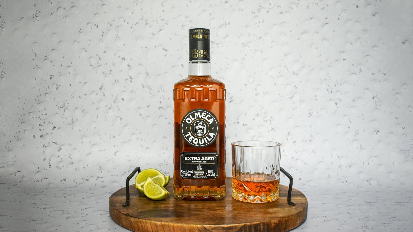 Botella de tequila, vaso y gajos de limón sobre una bandeja de madera.